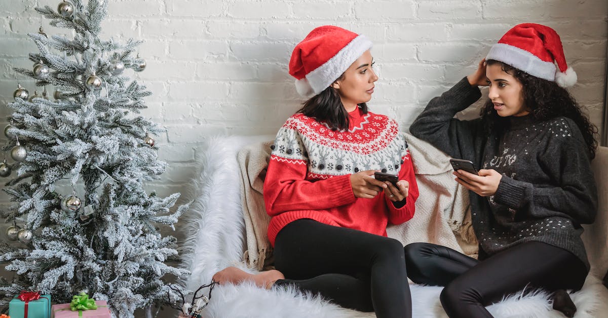 How to insert emoticons in the chat box? - Multiracial young friends in warm sweaters with mobile phone chatting near Christmas tree decorated with sparkling silver baubles