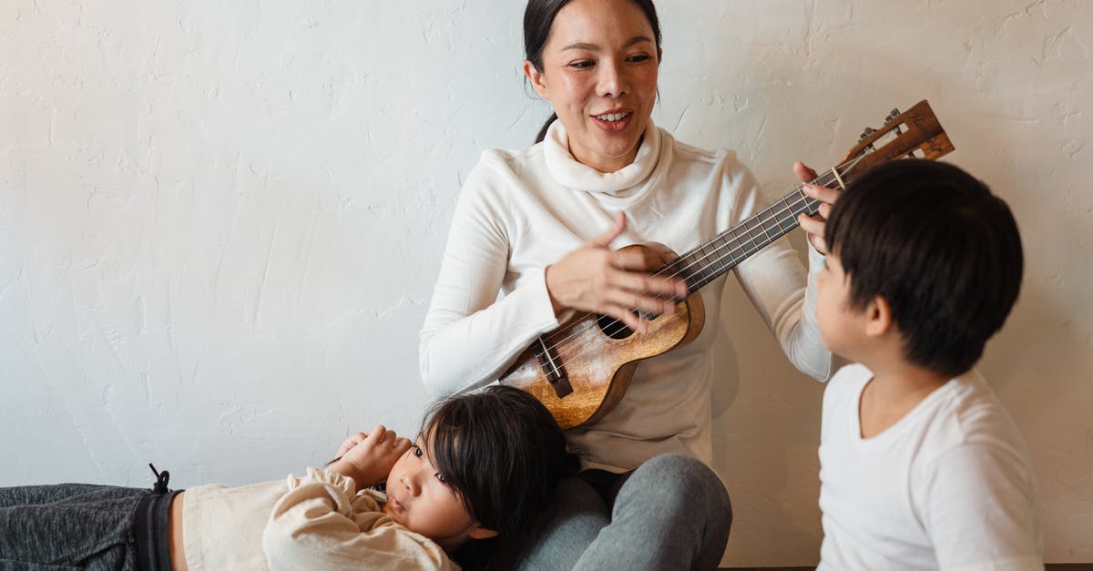 How to perform a flat spin? - Cheerful slim female musician playing acoustic ukulele while sitting with crossed legs in apartment near charming sons in casual wear