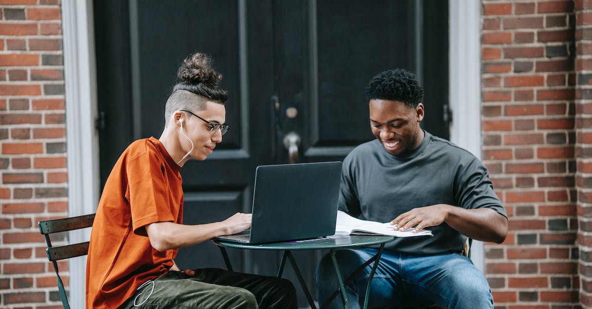 How to plan a good street layout - Positive young diverse male colleagues in casual clothes smiling and using laptop while working remotely on project sitting at table in street cafe