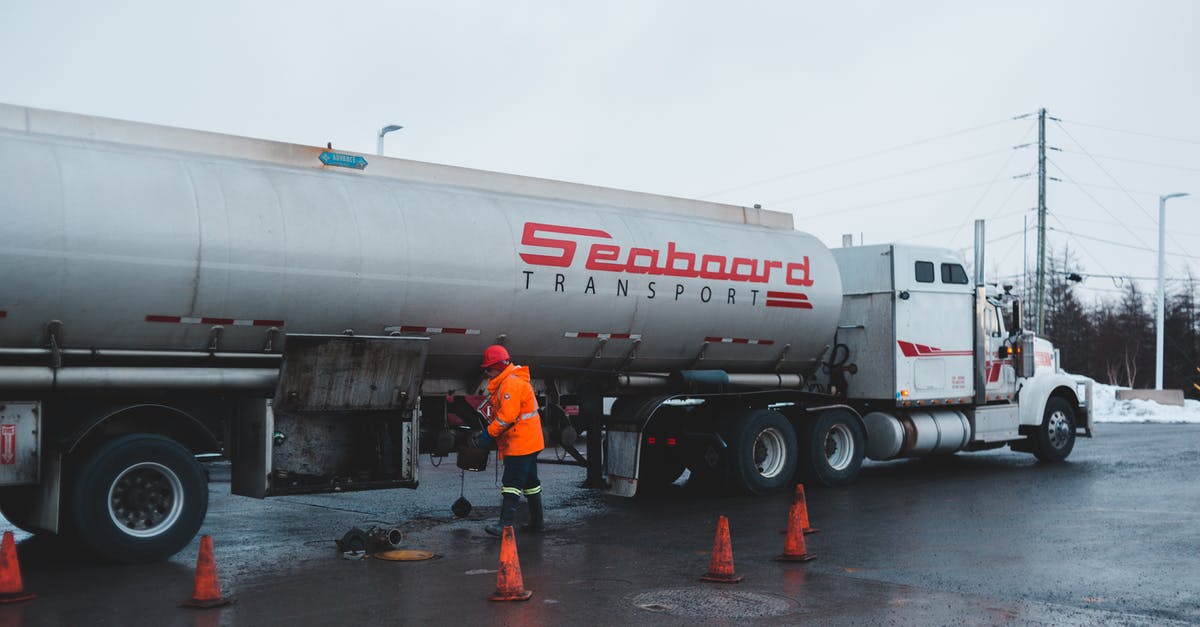 How to reload/refuel maintenance trailers? - Back view of anonymous male worker in bright uniform and helmet cleaning oil truck vehicle while standing on asphalt roadway surrounded by barriers under white sky in town