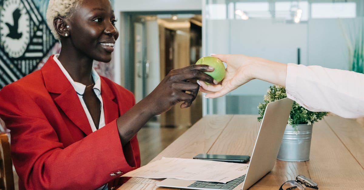 How to take away only one of multiple items [duplicate] - Side view of cheerful African American businesswoman in stylish clothes sitting at wooden table with netbook and documents and taking green apple from coworker in modern workspace
