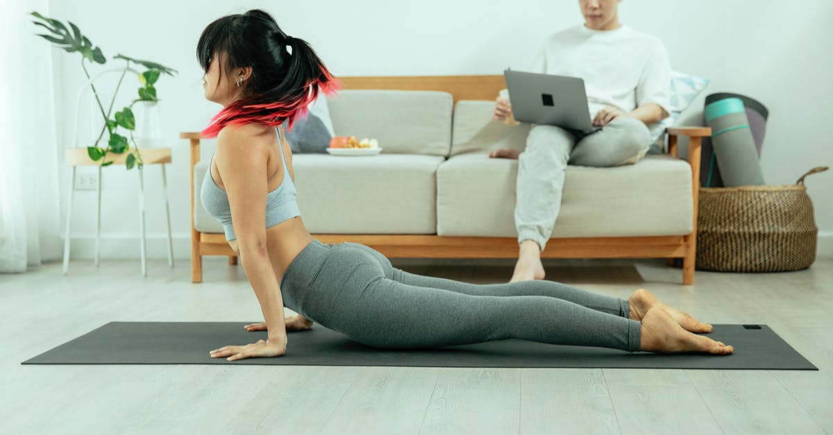 How to use those soul stones? - Side view young slim female wearing gray top and leggings stretching in Cobra while practicing Bhujangasana yoga pose on mat in room while man sitting on sofa browsing laptop