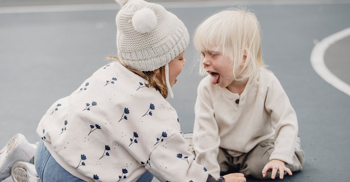 I have little lag on Offline Games - Side view of cute little girl with hat and playful boy showing tongue sitting on sports ground while having fun against blurred background