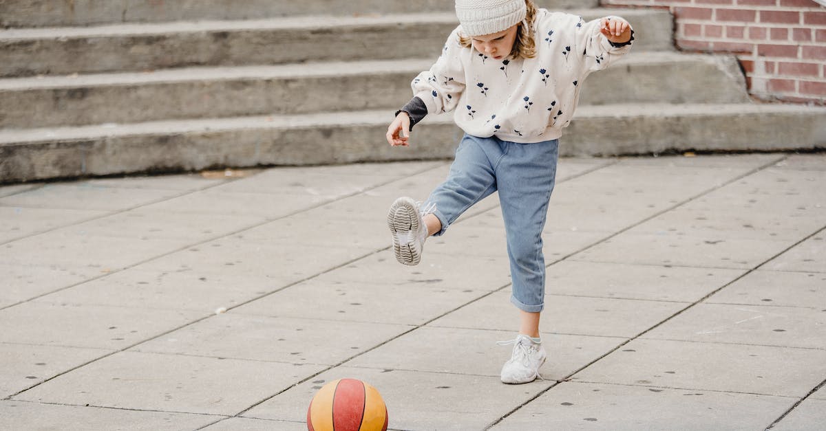 In all games, shadows have small rectangles that flicker - Full body of focused little girl in jeans white jumper and hat kicking colorful ball on street In all games, shadows have small rectangles that flicker - Full body of focused little girl in jeans white jumper and hat kicking colorful ball on street
