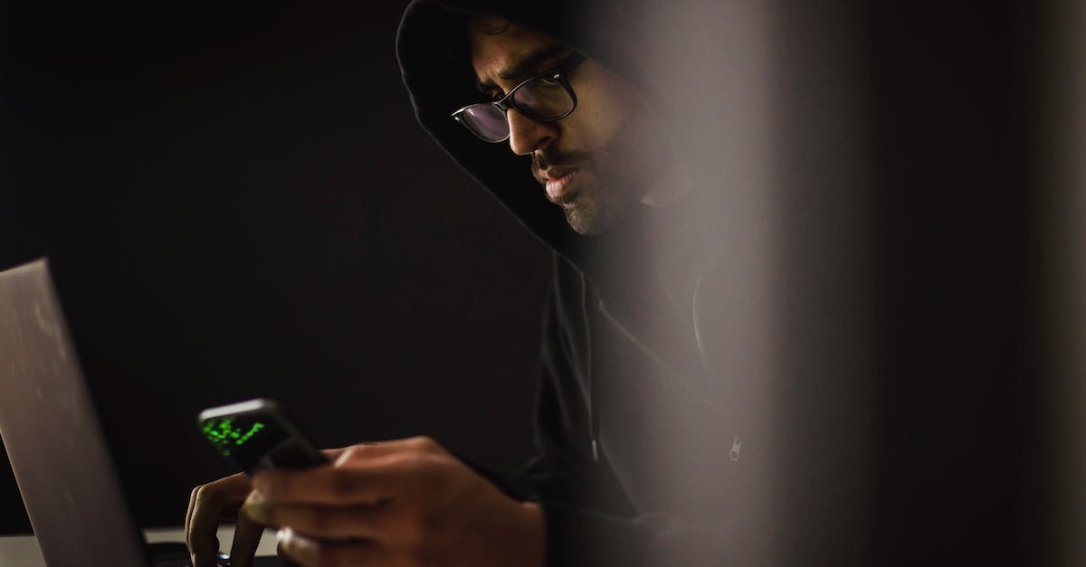 Is it possible to be immune to dark attacks? - Crop ethnic hacker with smartphone typing on laptop in darkness