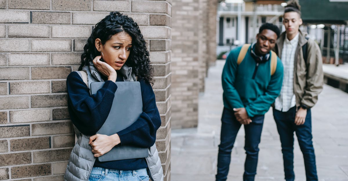 Is it possible to hide your backpack or helmet? - Latin American woman leaning on brick wall while hiding from multiracial classmates in college