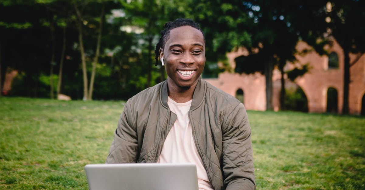 Is Quiet connected? - Positive African American male in casual clothes with wireless earbuds using laptop and smiling while sitting on green lawn and enjoying free time in quiet park Is Quiet connected? - Positive African American male in casual clothes with wireless earbuds using laptop and smiling while sitting on green lawn and enjoying free time in quiet park