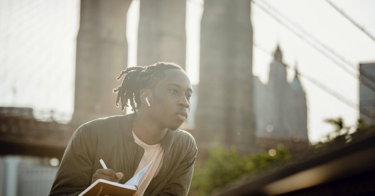 Is there a story behind the Doom Guy dolls? - Thoughtful young black man with dreadlocks listening to audio story in wireless earphones and writing down information in notebook while leaning on fence on city promenade
