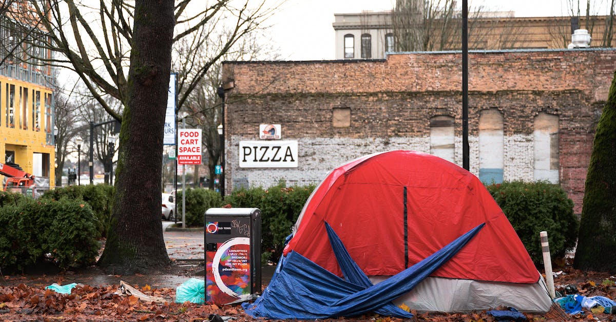 Is there a way to get rid of dirt patches? - Red tent for homeless people placed near asphalt road on street with tree and buildings in town against cloudless sky