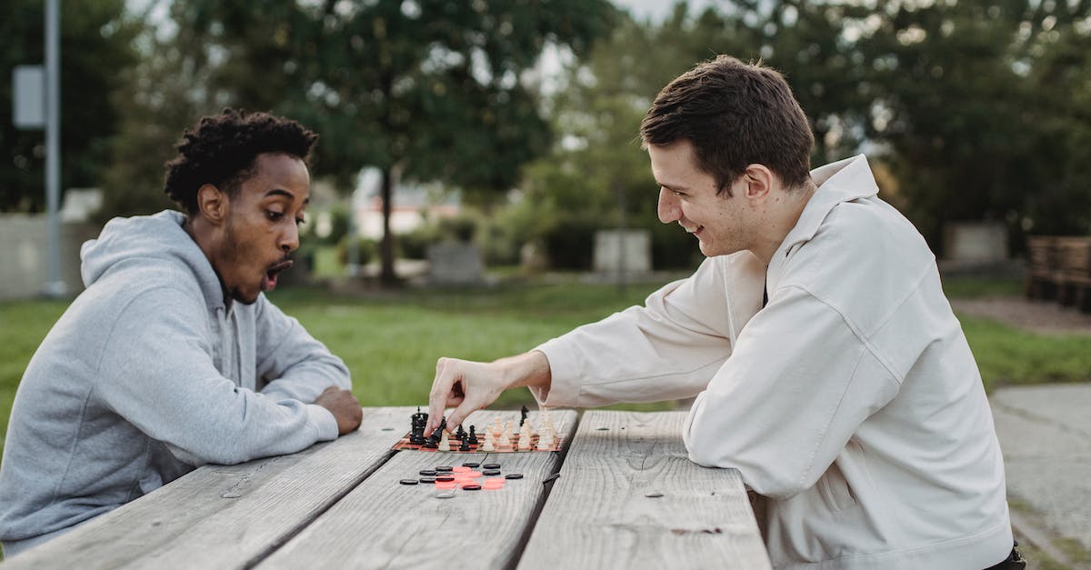 Is there a way to play the Battle Maison with friends over online? - Side view of smiling young multiracial male friends sitting at wooden table in city park and enjoying interesting chess game