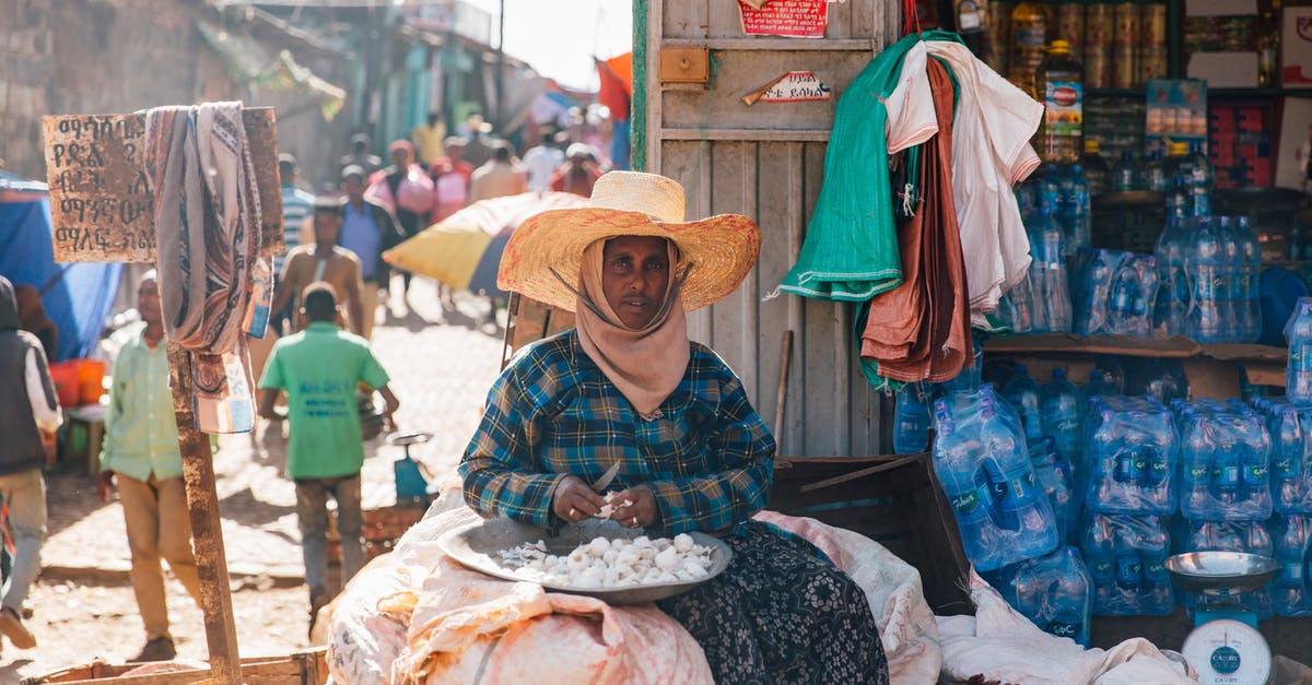 Is there any benefit to sending out a Bean Bottle from Poké Pelago? - Ethnic male in national clothes and hat looking at camera while sitting on sack with knife and plate full of garlic near product stall and people walking on old street