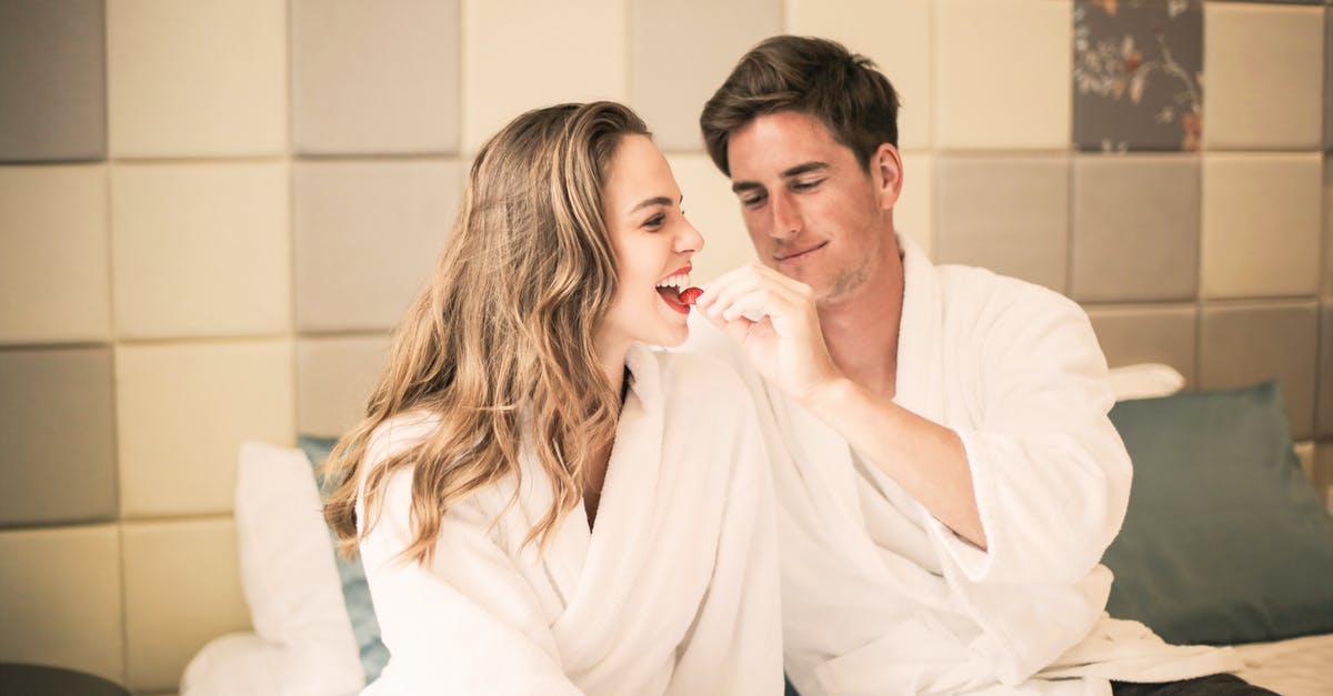 Is there any difference between a brahmin feed trough and a bathtub? - Delighted couple in bathrobes sitting on comfortable bed in cozy bedroom and feeding each other with delicious strawberry while relaxing at home