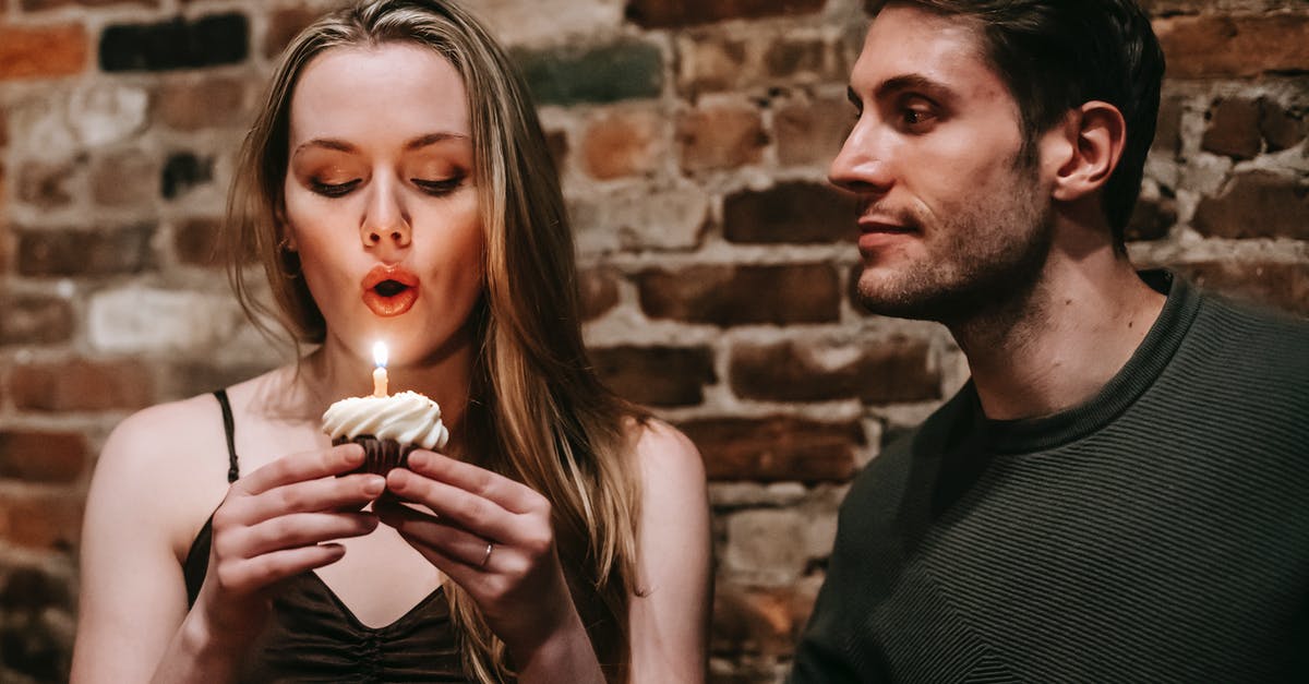 Is there any reasons not to make friends? - Young couple in elegant outfits in restaurant while blowing in candle on small cupcake and celebrating birthday near brick wall
