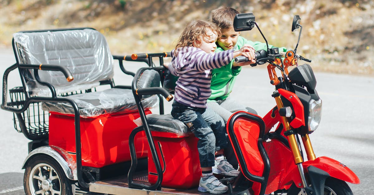 Is there any way to help Celina? - Adorable child helping little brother to ride modern red trike motorcycle on asphalt road on sunny day