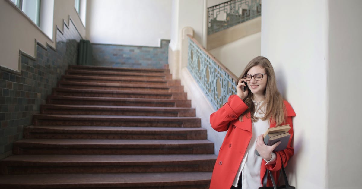 Is using the break room the only way to get some rest? - Cheerful female student standing with books and handbag in university hallway near staircase and speaking on mobile phone with closed eyes while relaxing after lesson and leaning on wall