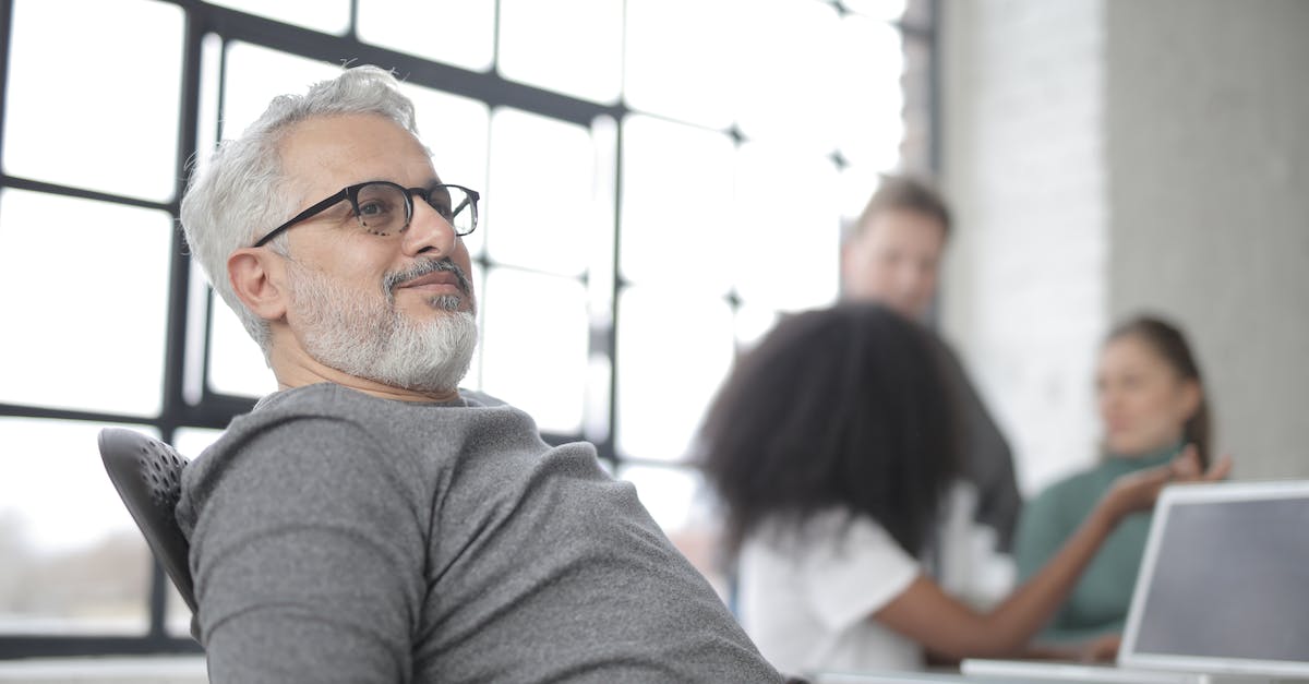 Leader died mid-conversation, will the ambassador come back next year? - Aged male boss wearing glasses resting in computer chair leaning back and with hands on armpads near desk with laptop while work on project at workplace with industrial interior with group of multiracial coworkers
