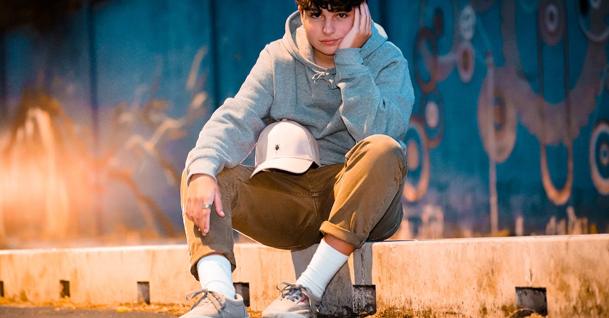 Level cap in Blitzball? - Ground level of crop stylish male adolescent in contemporary wear with cap sitting on barrier leaning on hand and looking at camera in evening Level cap in Blitzball? - Ground level of crop stylish male adolescent in contemporary wear with cap sitting on barrier leaning on hand and looking at camera in evening