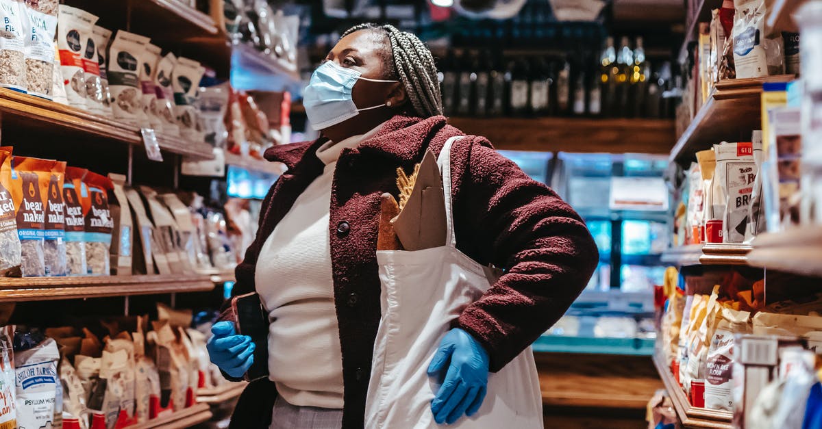 Logging in on a different phone - African American female buyer with shopping bag and smartphone in protective mask and gloves selecting products in supermarket