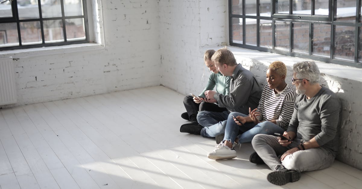 Logging in on a different phone - From above of diverse colleagues in casual wear browsing internet on cellphones while sitting with crossed legs near big fenced window in afternoon
