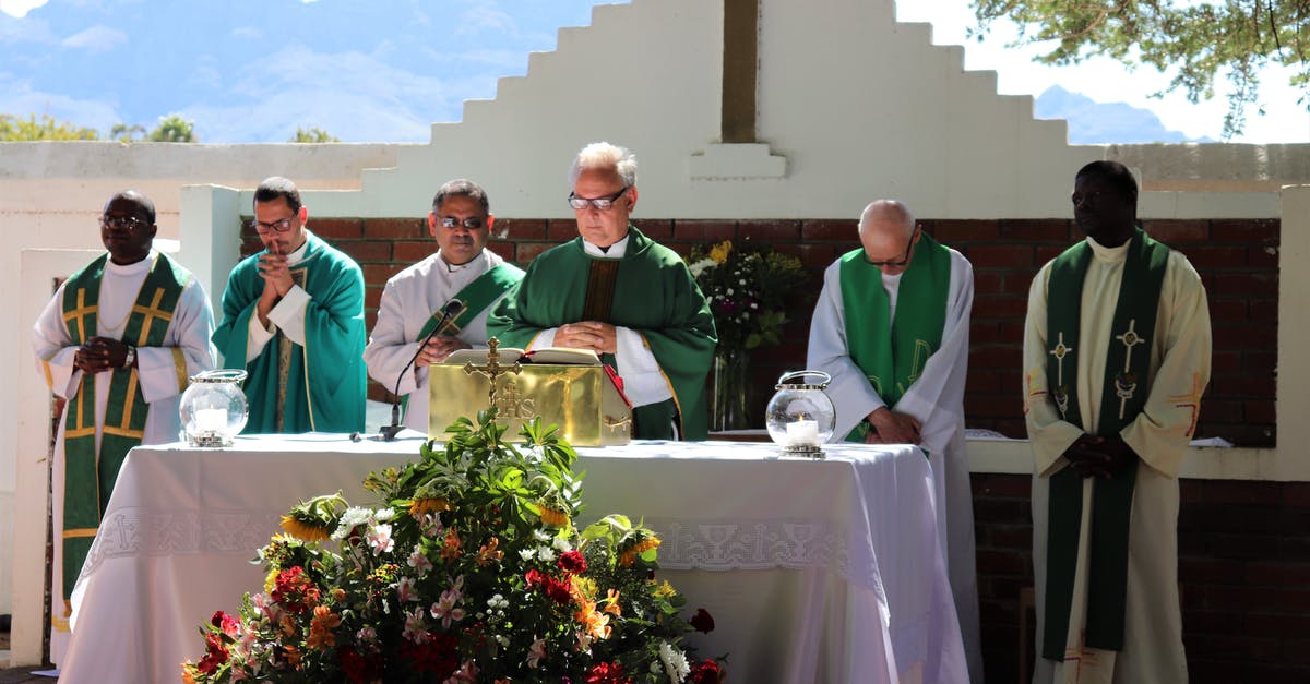 Mass Effect 3 Motivational Speech - Elderly priest in eyeglasses and catholic vestment standing with prayer hands near table with Bible and multiethnic deacons during liturgy process behind mountains in sunlight