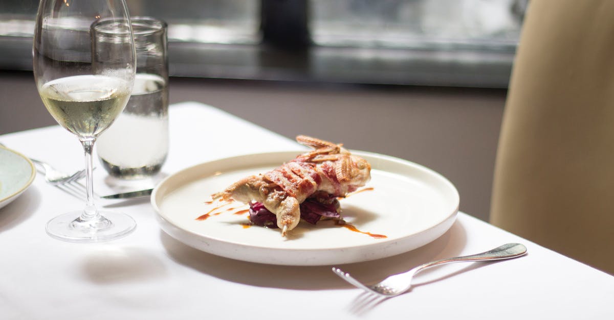 Missing my last bird in Botanicula, at restaurant - Tablecloth on table with delicious plate with small roasted bird covered in bacon with sauce near glasses with wine and water near forks and knife in light restaurant near chair