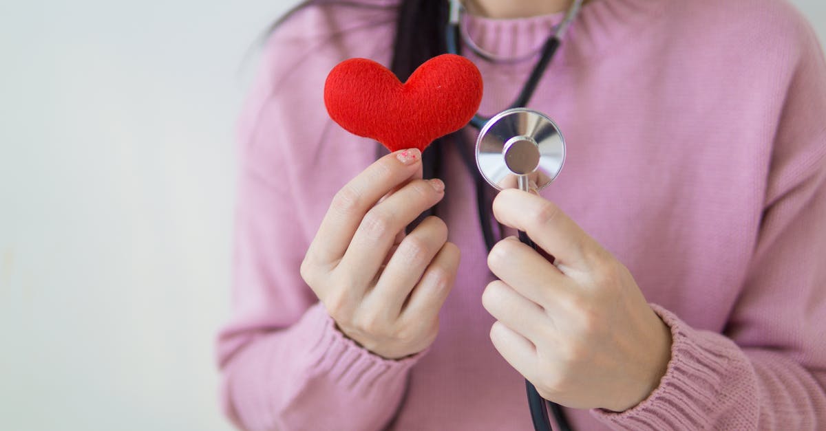 Mob Health Check [duplicate] - Unrecognizable female in pink sweater with stethoscope on neck standing on white background with red heart in hand in daylight
