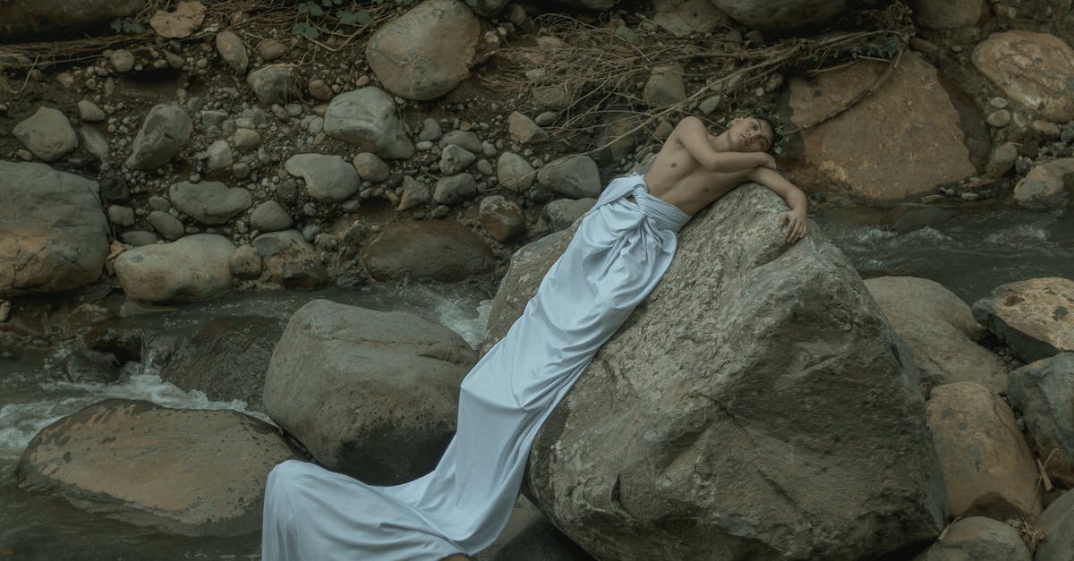 Mysterious White Dogtag - Male in long white sheet resting on heavy rough stone near splashing stream Mysterious White Dogtag - Male in long white sheet resting on heavy rough stone near splashing stream