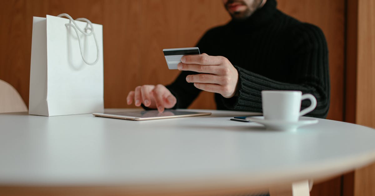 Name for games that have abusive in app purchases - Crop focused bearded young man using tablet while doing online purchase with credit card sitting at table in modern cafe with coffee cup and shopping bag