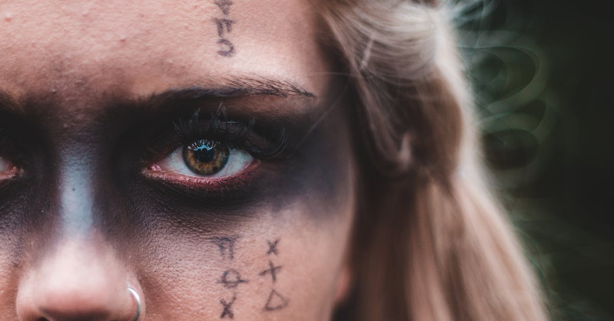 Piercing or Critical For Magic Attacks? - Closeup crop blond with black painted mask on eyes and symbols on skin looking at camera Piercing or Critical For Magic Attacks? - Closeup crop blond with black painted mask on eyes and symbols on skin looking at camera