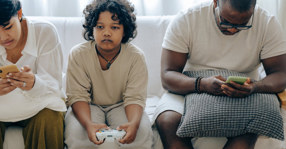 playing Digital game online with my family member > - Focused ethnic child playing video game with joystick sitting on sofa between divers parents browsing smartphones playing Digital game online with my family member > - Focused ethnic child playing video game with joystick sitting on sofa between divers parents browsing smartphones