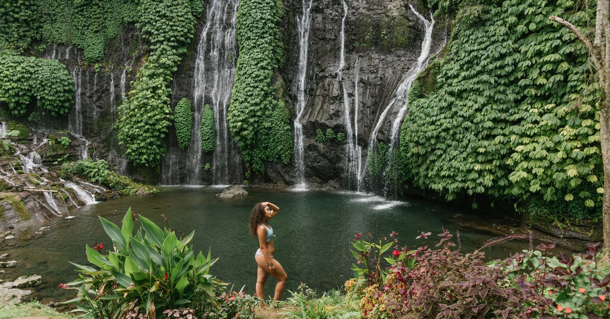 Possible to recreate lakes? - High angle of peaceful young woman in bikini touching hair while standing between tropical plants against waterfall flowing into pond