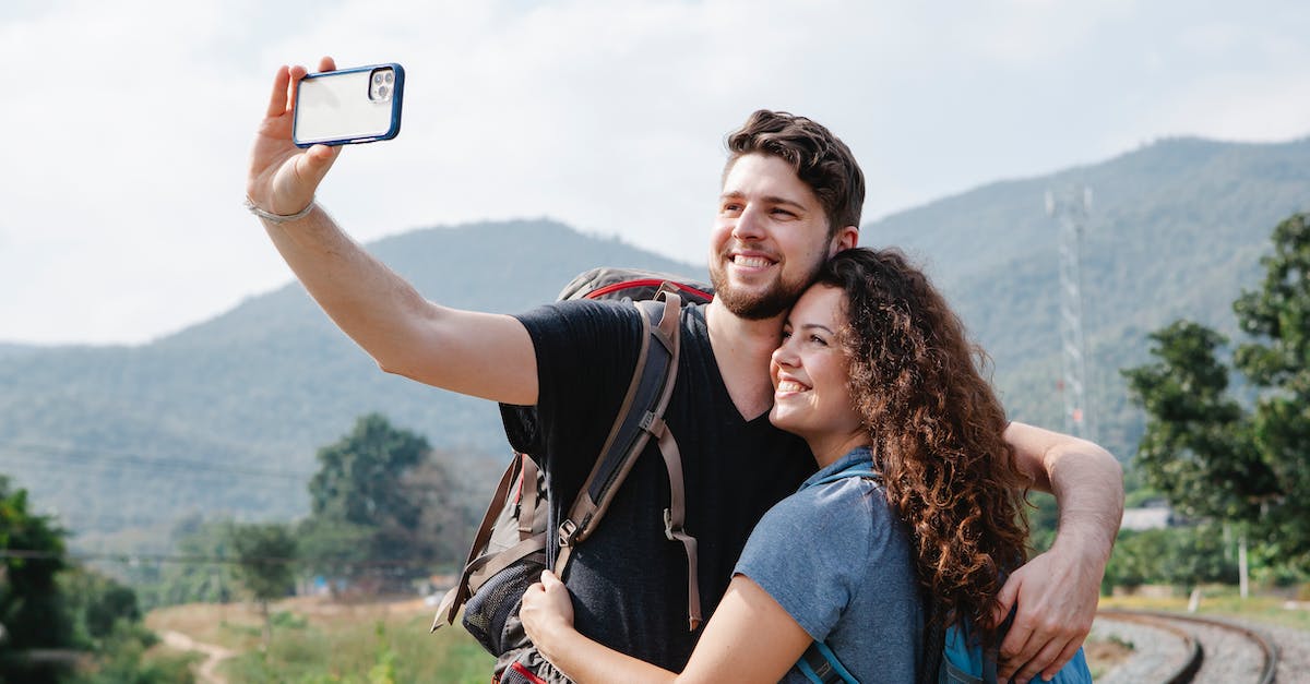 Railroad Tycoon II screen resolution - Cheerful couple of tourists in casual clothes with hiking backpacks hugging and taking selfie on smartphone in nature against green hills in daytime