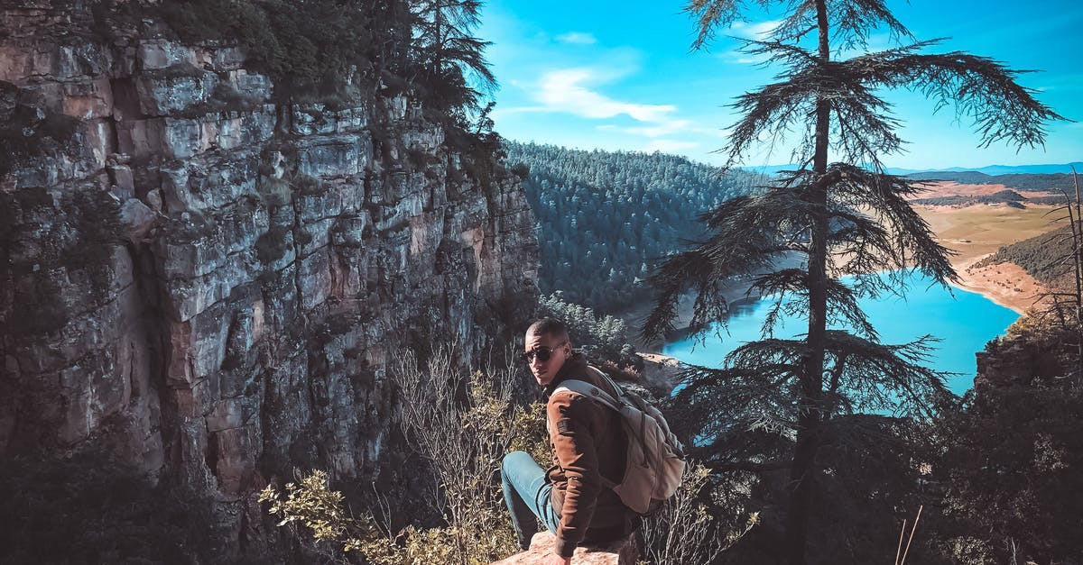 Rapidfire Cannon on Assassins, concern over range - Back view full length male wearing casual clothes sitting on edge of stony cliff against rough mountains and calm blue river while spending time in amazing highlands on clear day