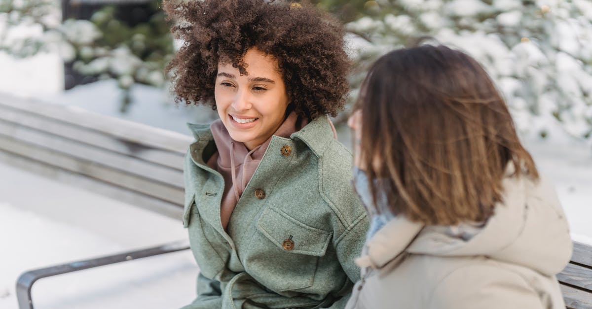 Removing freeze time forever - High angle of positive African American female with curly hair sitting on bench near girlfriend while spending time in winter park Removing freeze time forever - High angle of positive African American female with curly hair sitting on bench near girlfriend while spending time in winter park