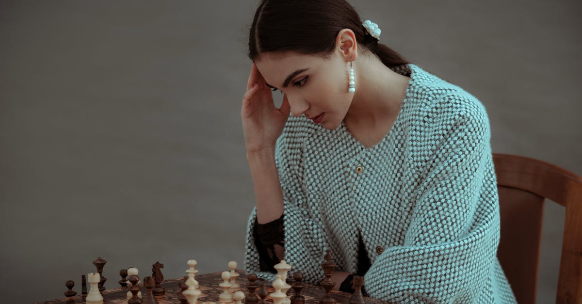 Shield enemies tactic - Concentrated ethnic female sitting on chair and touching head while playing chess against blurred gray background Shield enemies tactic - Concentrated ethnic female sitting on chair and touching head while playing chess against blurred gray background