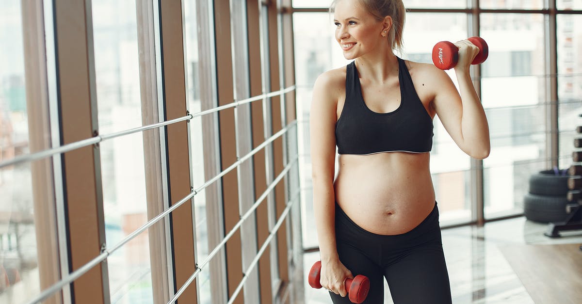 Should I wait to reclaim a gym? - Young pregnant female in sportswear smiling and looking away while standing near glass wall in modern gym and training with red dumbbells Should I wait to reclaim a gym? - Young pregnant female in sportswear smiling and looking away while standing near glass wall in modern gym and training with red dumbbells