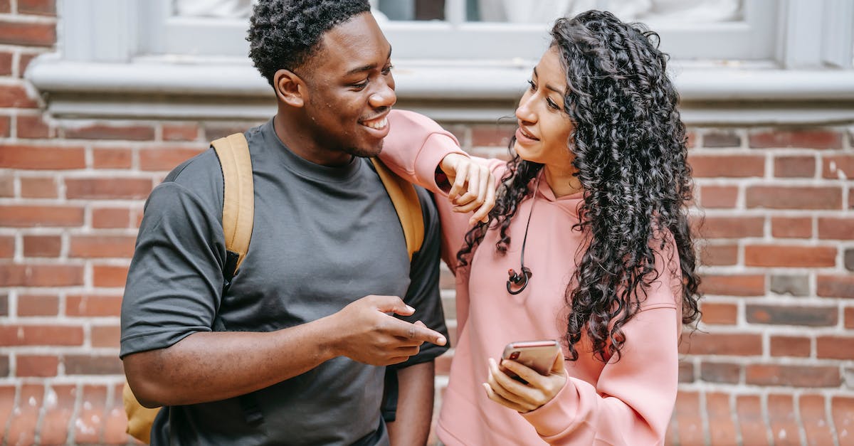 Show radiuses of other buildings - Positive young ethnic female with long curly hair standing on street with arm on shoulder of cheerful black boyfriend and showing funny video on smartphone