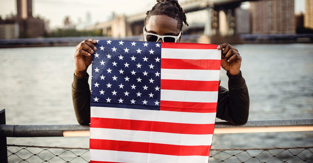 Show scoreboard belowname without a value - Ethnic male in casual clothes and sunglasses standing on embankment of city river while leaning on fence showing national flag of United States of America