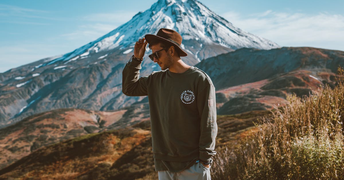 Sightseeing: Tollifer's Last Stand - Young stylish man in hat and modern sunglasses standing on hill with hand in pocket against high snowy mountain