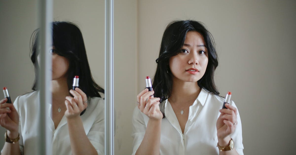 Skyrim Fortify Smithing Option does not appear - Young Asian woman looking at camera and selecting between lipsticks while standing near mirror during makeup application at home