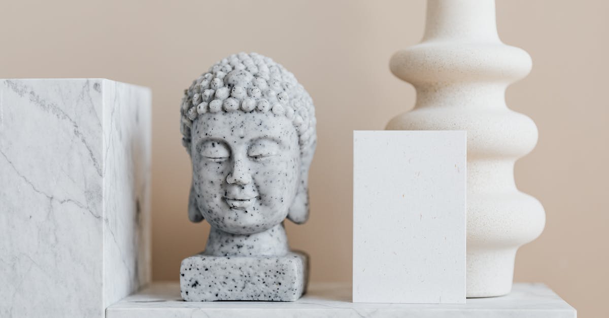 Stone heads in Lhe a Rhemen - Granite bust of Buddha placed near white ceramic vase of creative geometric shape and blank card on white marble shelf against beige wall