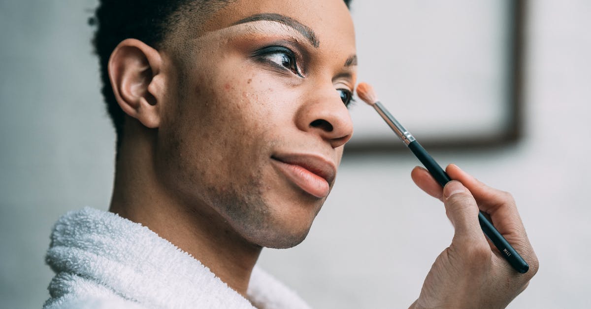 Strange lighting anomaly - Side view of queer African American male in white bathrobe applying eyeshadow with cosmetic brush in light room at home Strange lighting anomaly - Side view of queer African American male in white bathrobe applying eyeshadow with cosmetic brush in light room at home