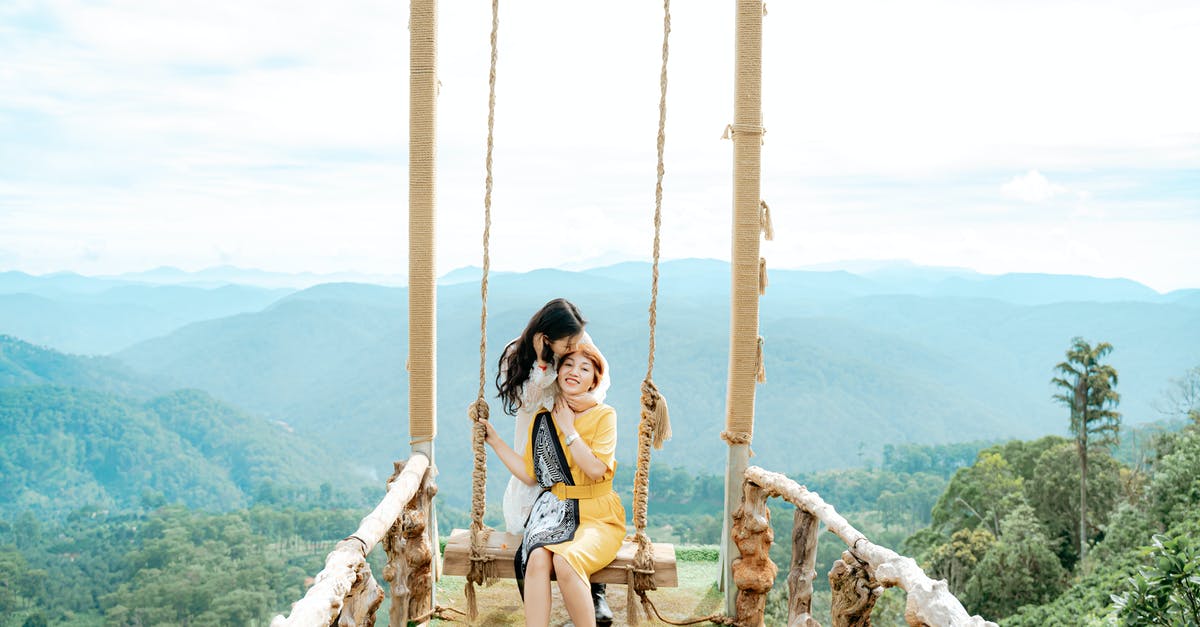 Stuck at swing bridge in Hunrath - Ethnic female traveler touching hair while kissing mom on swing against misty mounts under cloudy sky Stuck at swing bridge in Hunrath - Ethnic female traveler touching hair while kissing mom on swing against misty mounts under cloudy sky
