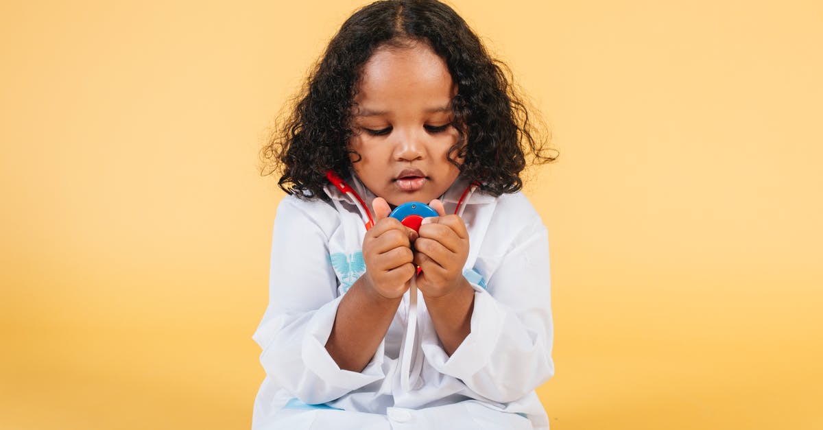 Stuck in color dungeon, one little key short - Thoughtful black girl with curly hair in white medical costume looking at toy stethoscope against yellow background in studio