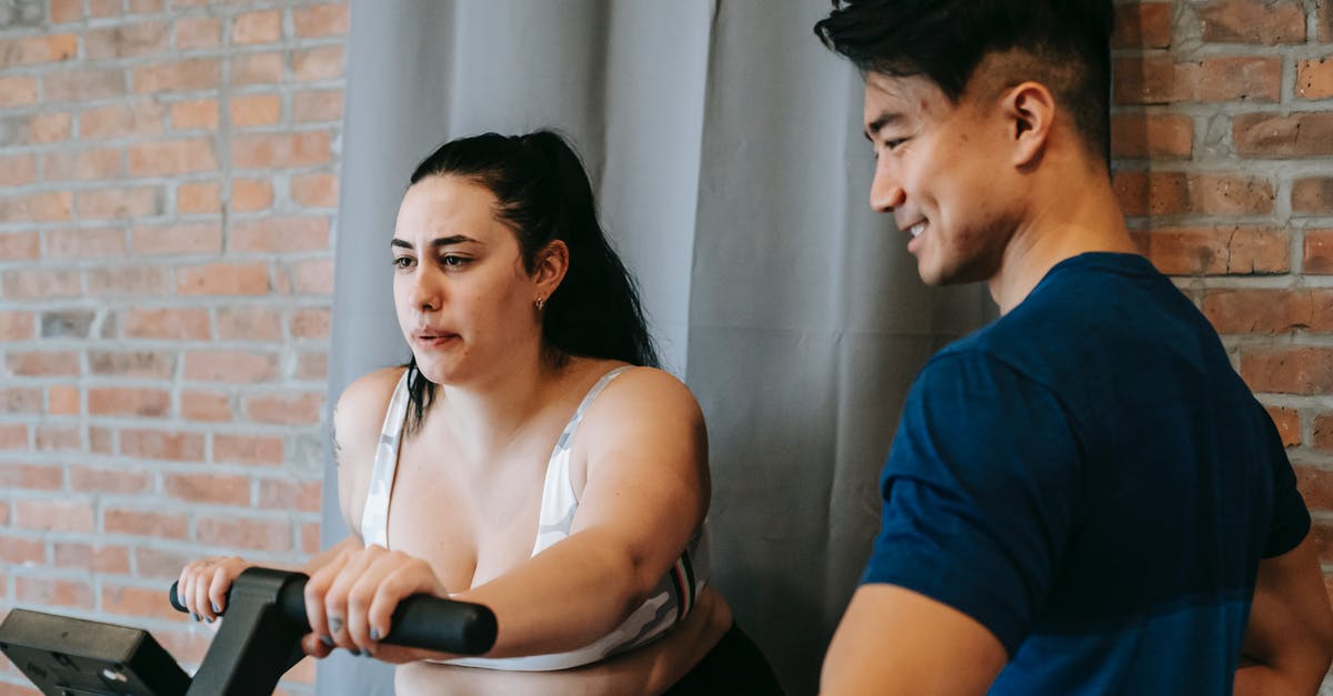 Training Mode: NPC on control option - Side view of positive young Asian male trainer supporting plus size female doing cardio workout on cycling machine in modern fitness center Training Mode: NPC on control option - Side view of positive young Asian male trainer supporting plus size female doing cardio workout on cycling machine in modern fitness center