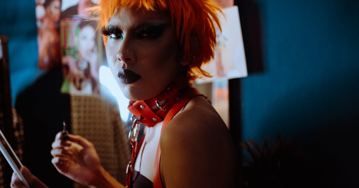 Unlocking the alternative side-quests? - Side view of serious transsexual ethnic male with eccentric makeup in orange wig and BDSM collar standing in dressing room and looking at camera
