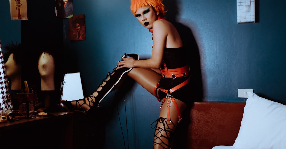 Unlocking the alternative side-quests? - Side view unemotional extravagant female with orange dyed hair and dark makeup wearing bodysuit and thigh high shoes putting leg on vanity table and looking at camera