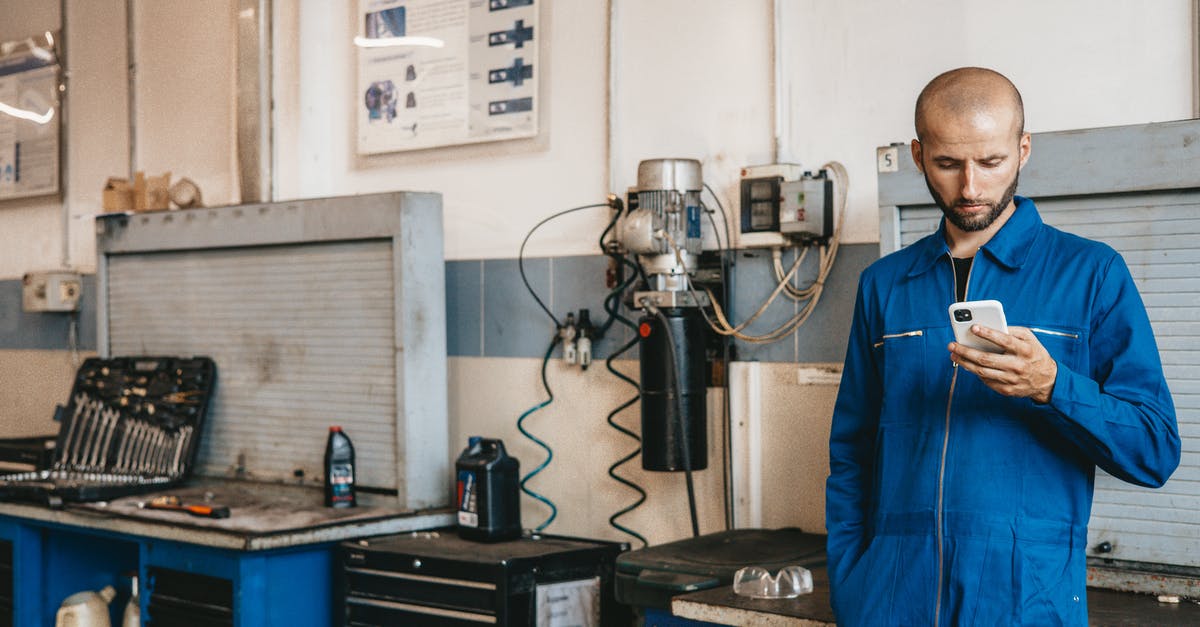 Use for upgraded tools - A Mechanic in Blue Coverall Using His Cellphone Inside The Workshop Use for upgraded tools - A Mechanic in Blue Coverall Using His Cellphone Inside The Workshop