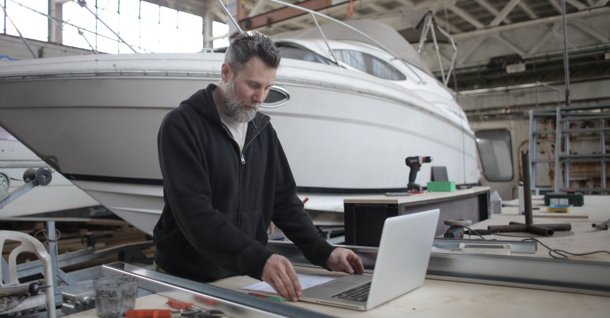 Using the collector on the rescue ship? - Thoughtful adult worker using laptop while working with metal parts near boat in workshop Using the collector on the rescue ship? - Thoughtful adult worker using laptop while working with metal parts near boat in workshop