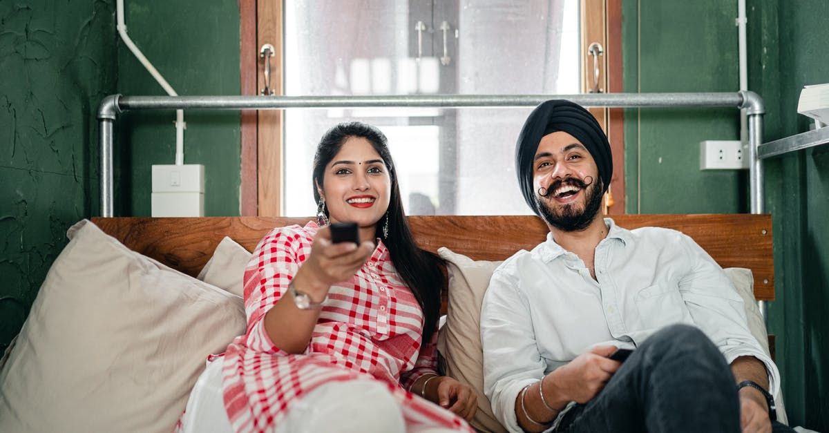 Watch won't show time - Cheerful wife with bindi on forehead wearing plaid tunic with white trousers using TV remote control for channel switching while lying on bed with laughing Sikh husband in turban with stylish beard and twisted mustache Watch won't show time - Cheerful wife with bindi on forehead wearing plaid tunic with white trousers using TV remote control for channel switching while lying on bed with laughing Sikh husband in turban with stylish beard and twisted mustache